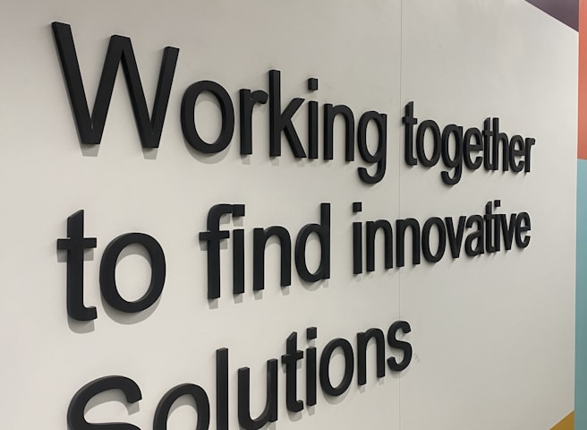 Black 3D text displays the phrase 'Working together to find innovative Solutions' against a geometric background with white, yellow, light blue, and dark blue areas.